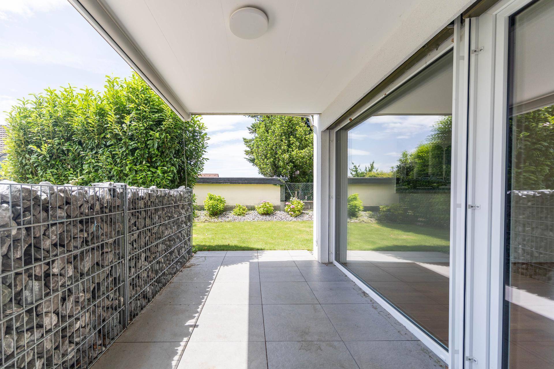 Überdachte Terrasse mit Fliesenboden und Gabionenwand, angrenzend an eine Rasenfläche.