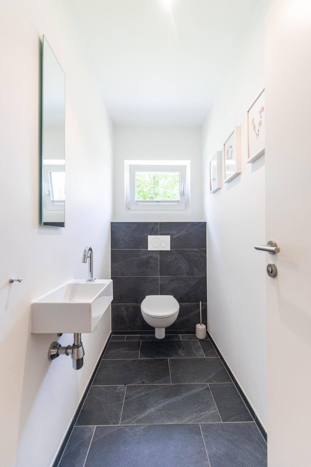Modernes Gäste-WC mit dunklen Fliesen, wandhängendem WC, kleinem Waschbecken und Fenster.