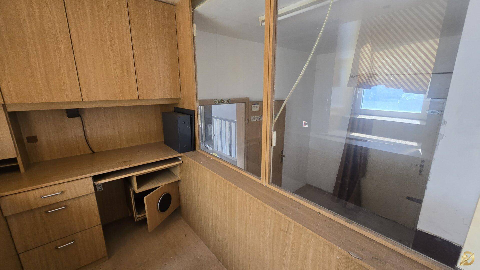 Detailansicht des Schreibtischbereichs im Büro mit Holzmöbeln und einem Fenster zu einem Nebenraum.
