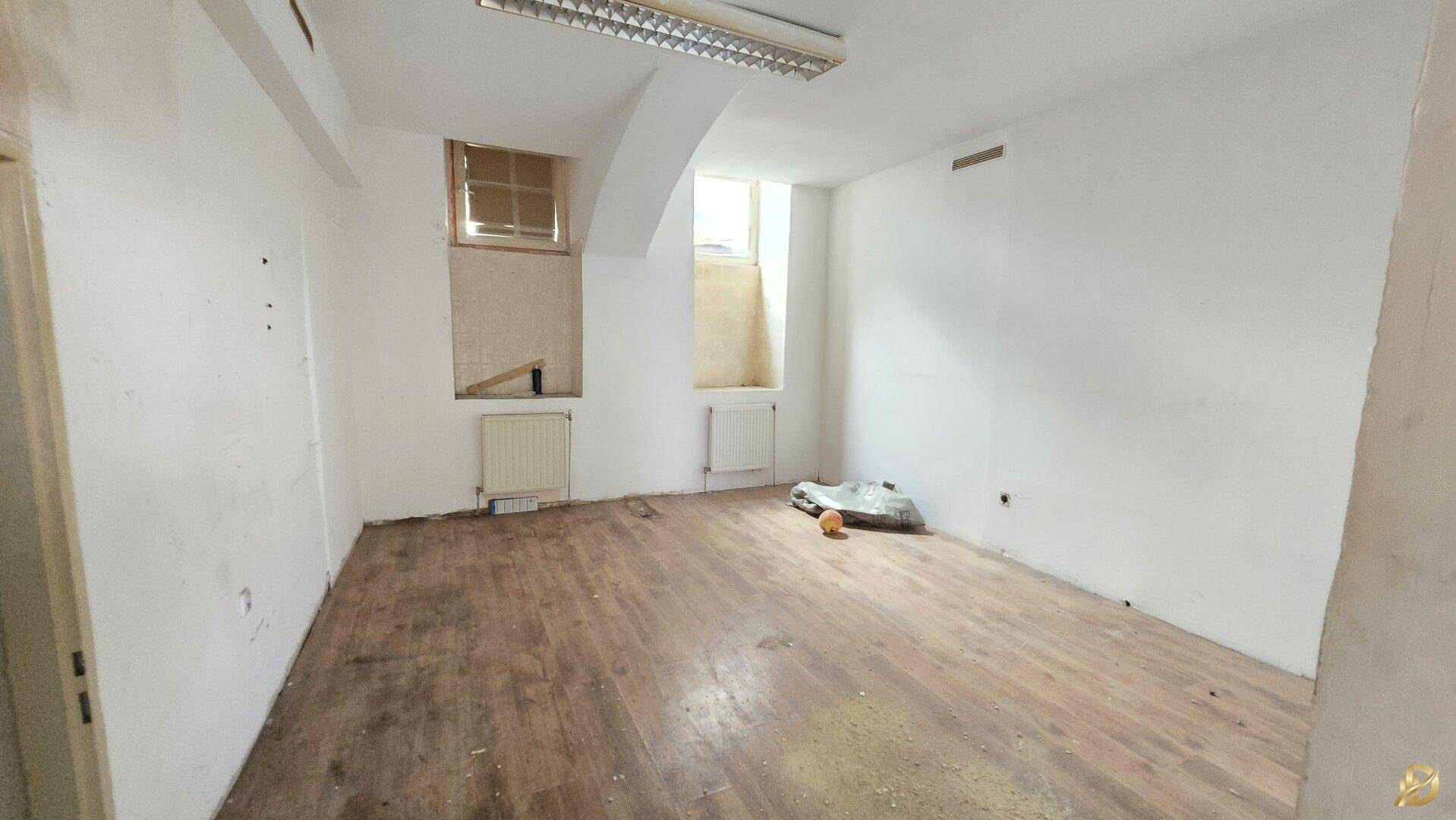 Leerer Büroraum mit Holzboden, weißen Wänden und zwei kleinen Fenstern, bereit zur Renovierung.