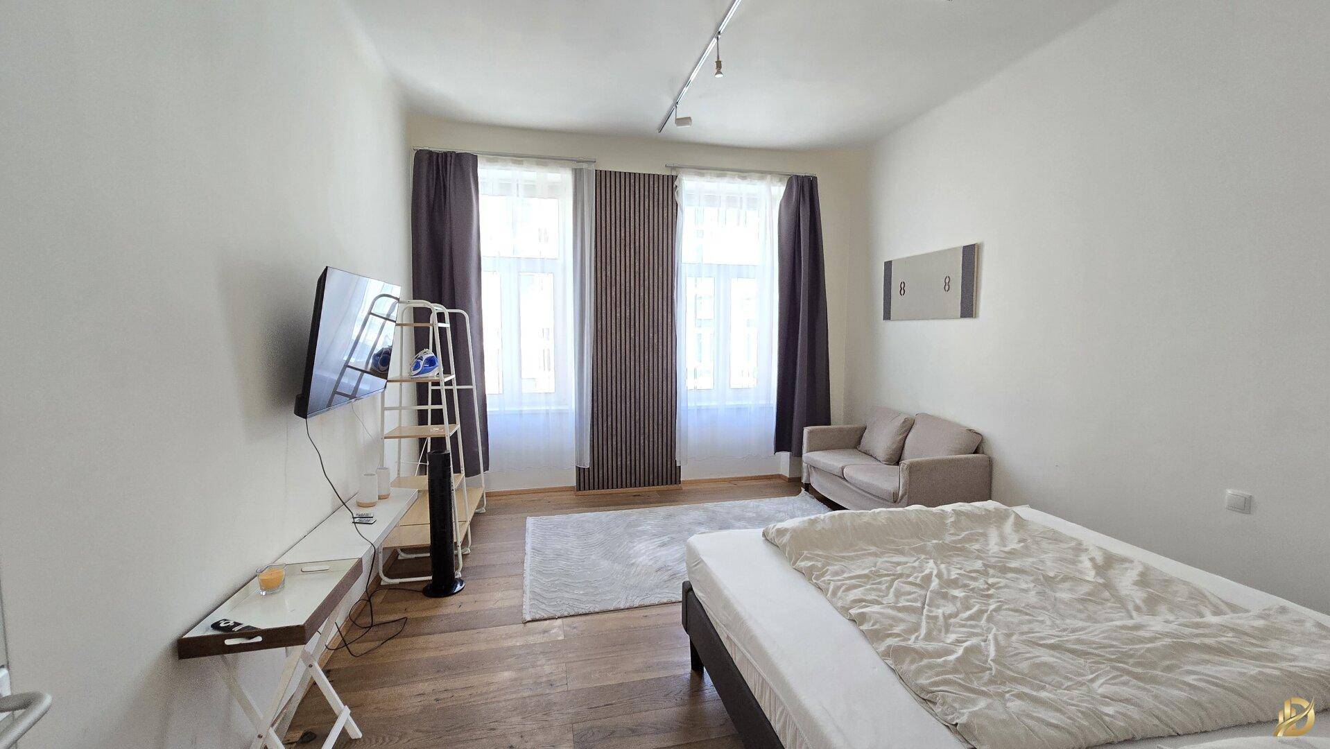 Geräumiges Schlafzimmer mit zwei Fenstern, einem Sofa und einem Fernseher an der Wand.