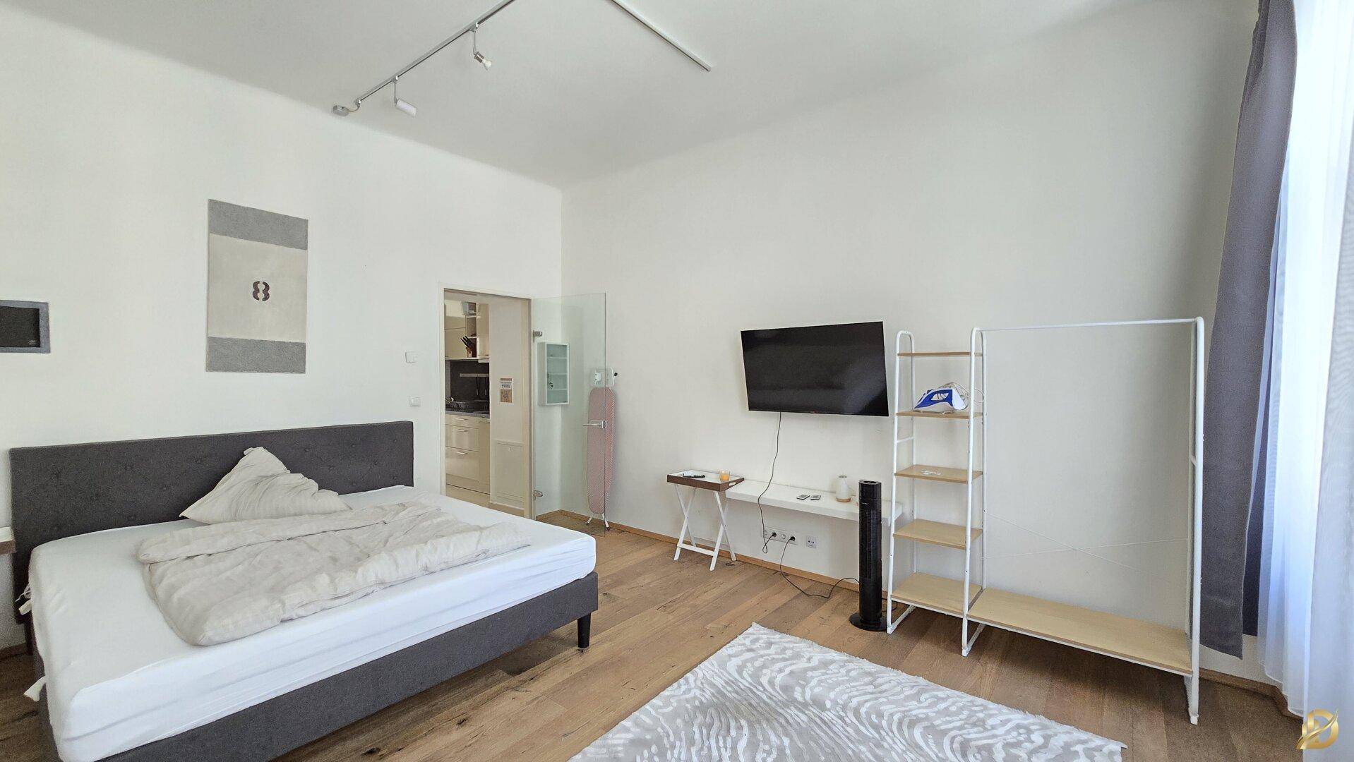Modern eingerichtetes Schlafzimmer mit Doppelbett, Wandfernseher und offenem Zugang zur Küche.