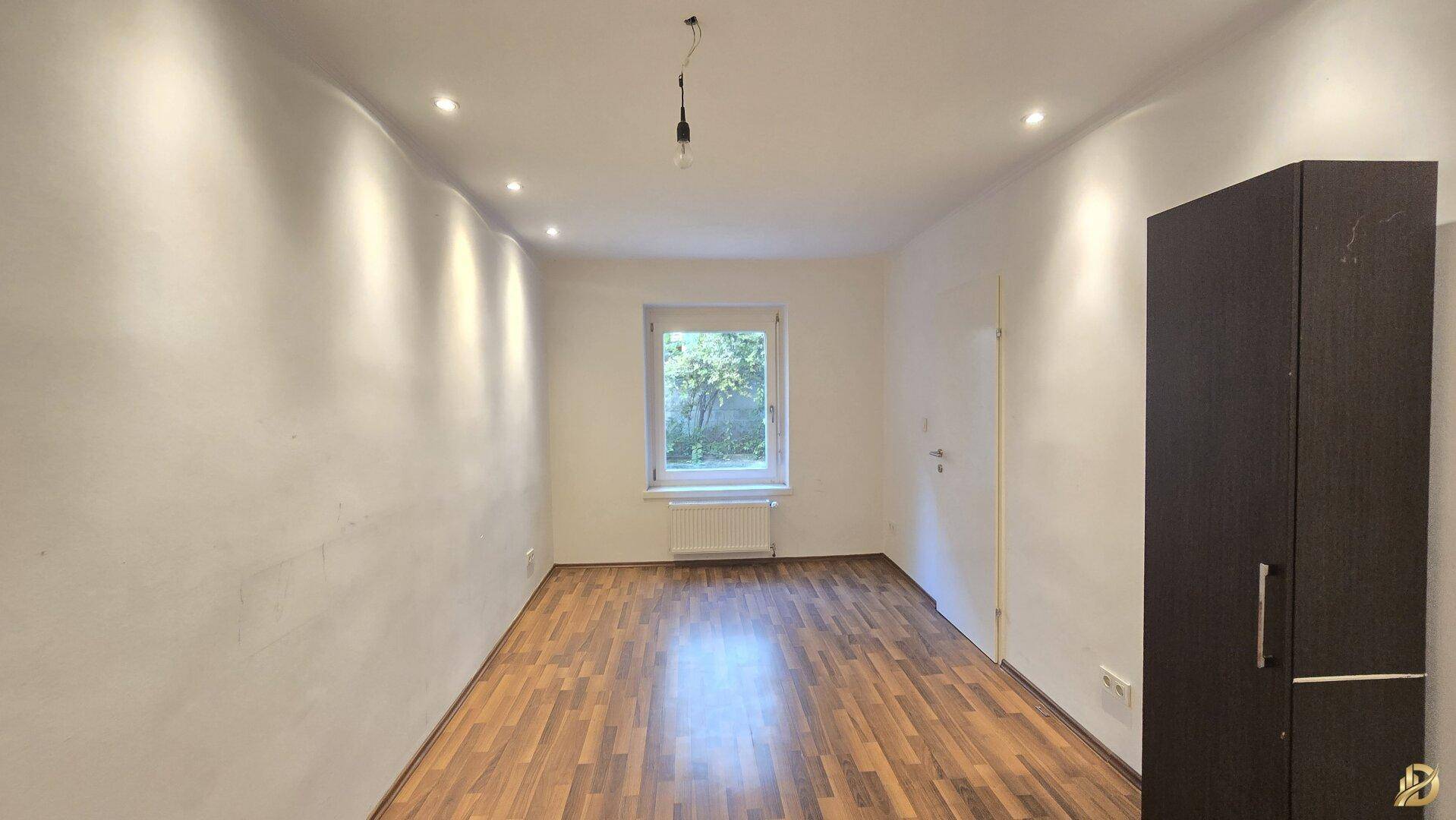 Helles Zimmer mit Parkettboden und einem Fenster mit Blick ins Grüne.