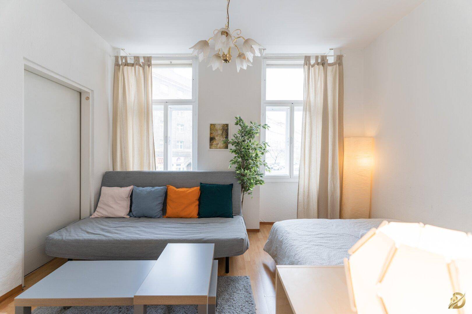 Helles Wohnzimmer mit Schlafsofa, Doppelbett und Holzboden, dekoriert mit Pflanzen und Kissen.