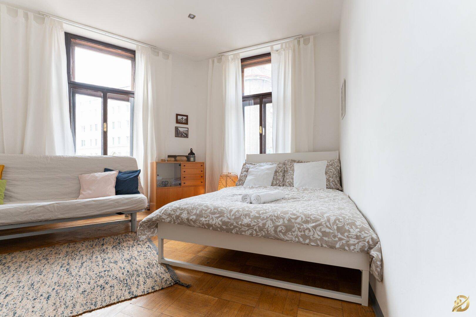 Gemütliches Wohnzimmer mit Schlafbereich, Holzboden und heller Einrichtung, gut beleuchtet durch Fenster.