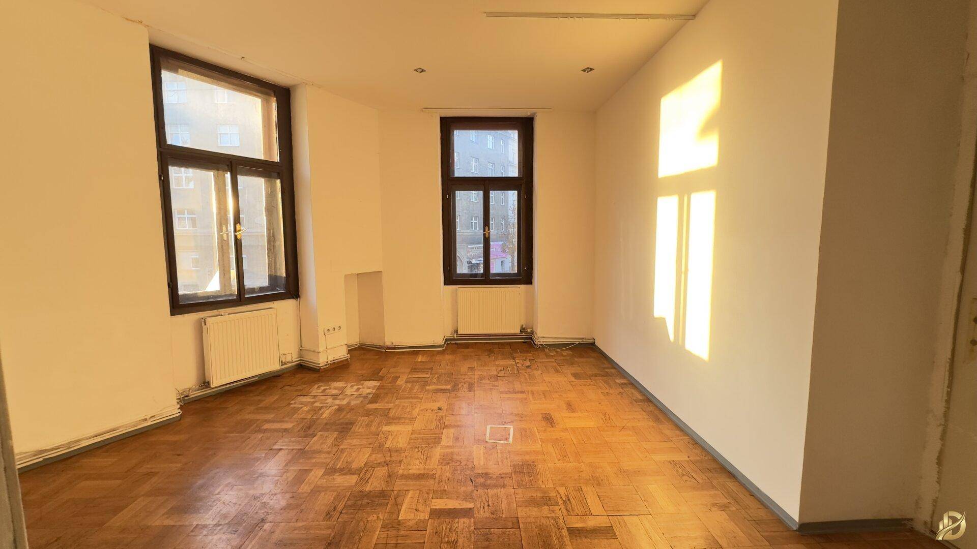 Helles Zimmer mit Fischgrätparkett und großen Fenstern, die viel Sonnenlicht hereinlassen.