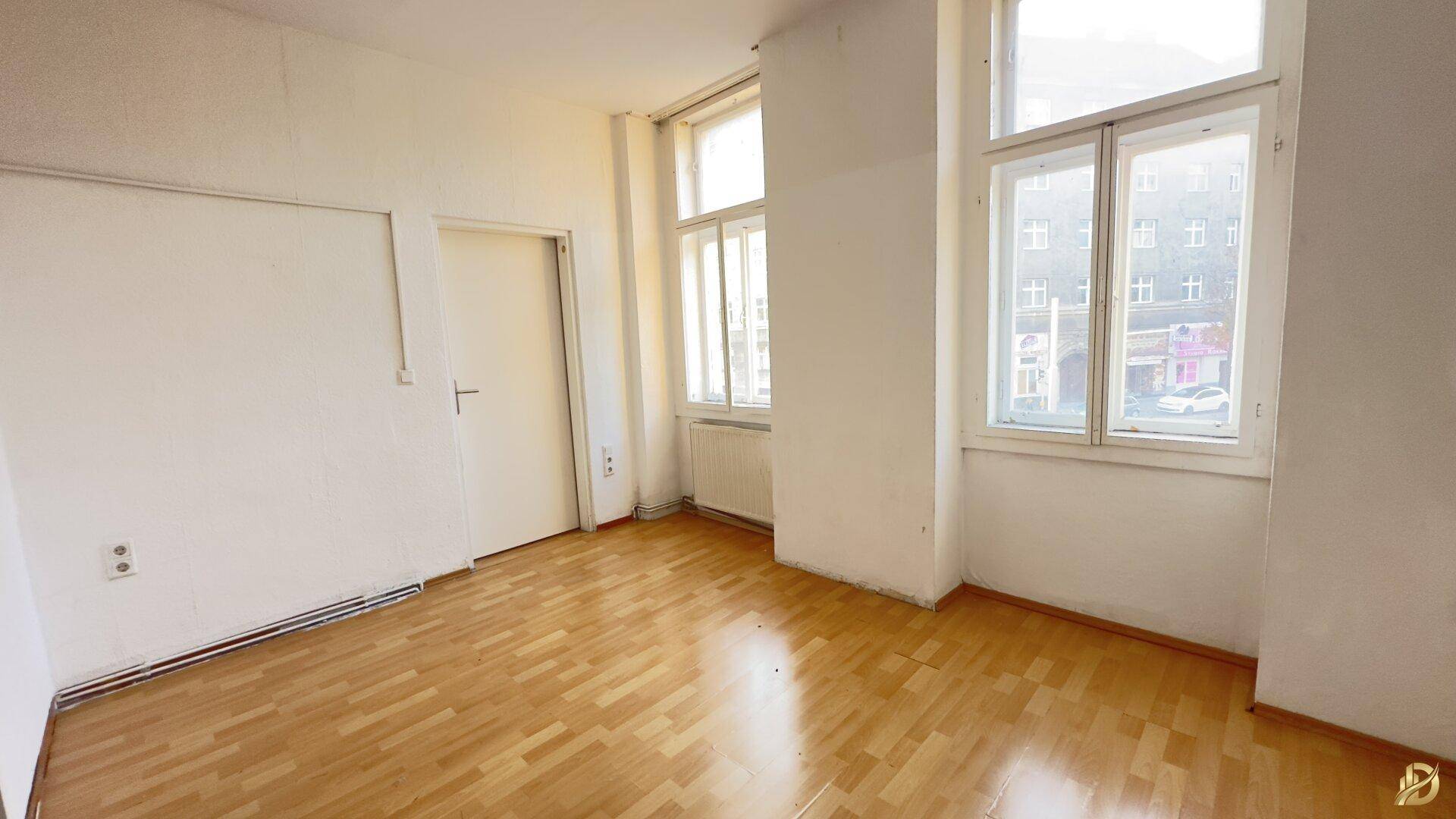 Leeres Zimmer mit Parkettboden und zwei großen Fenstern, die viel Tageslicht hereinlassen.