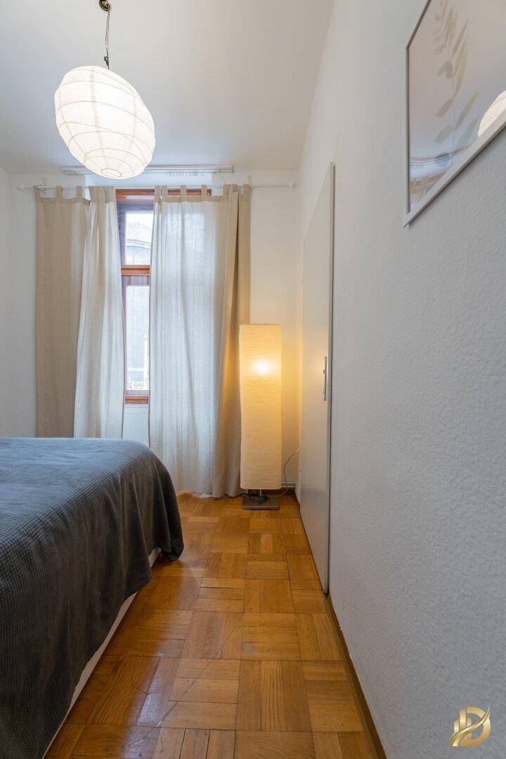 Schlafzimmer mit Doppelbett, Parkettboden und einem Fenster mit hellen Vorhängen.