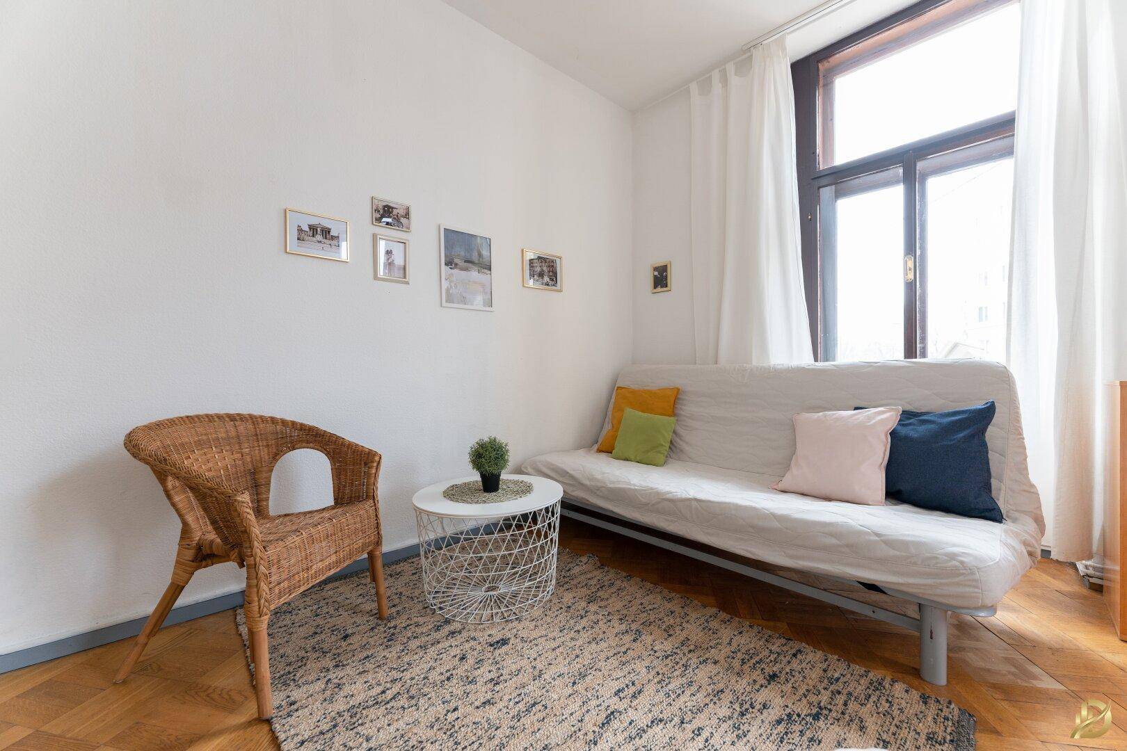 Gemütliches Wohnzimmer mit Schlafsofa, Korbsessel und Holzboden, gut beleuchtet.