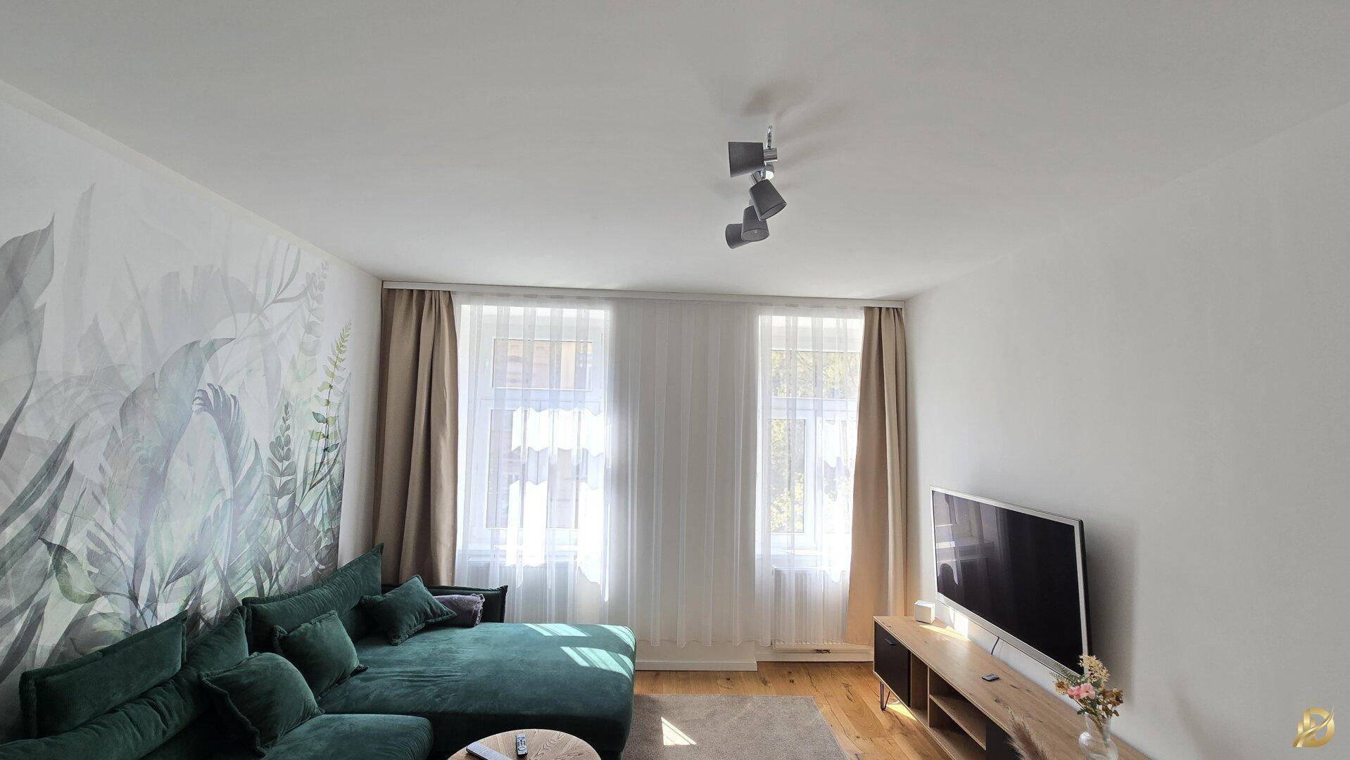 Gemütliches Wohnzimmer mit grünem Ecksofa, Fernseher und großen Fenstern, hell und modern gestaltet.