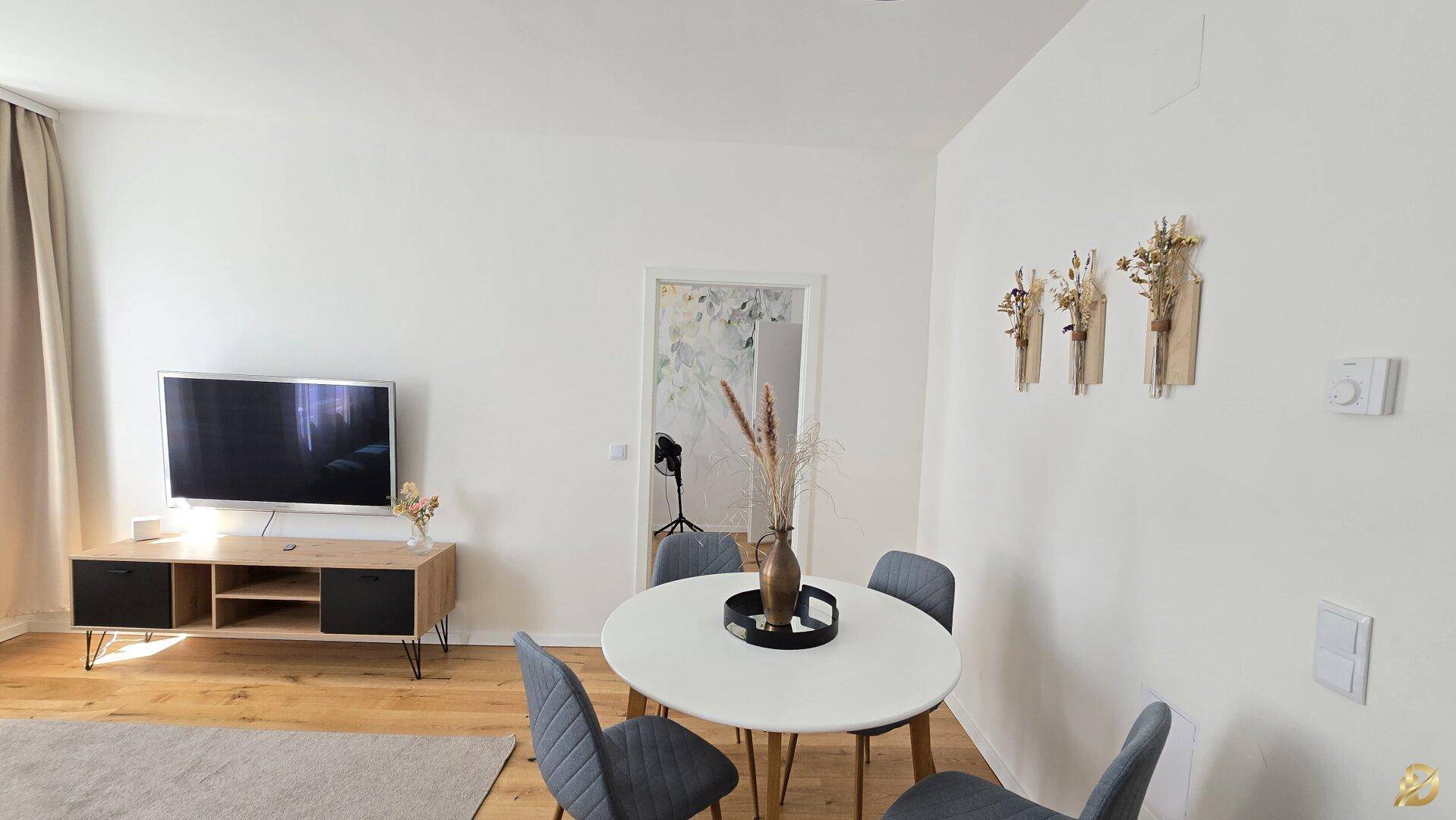Heller Essbereich mit rundem Tisch und Stühlen, angrenzend an den Wohnbereich mit Fernseher.