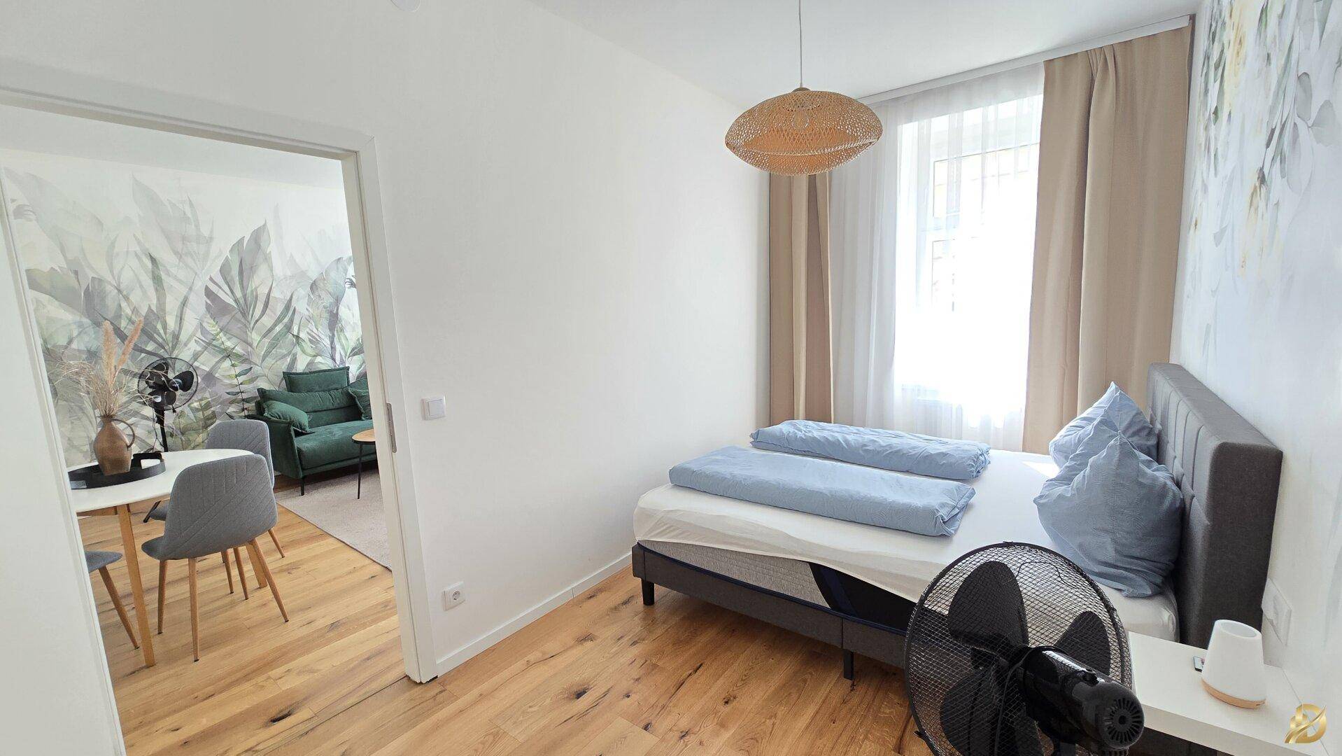 Schlafzimmer mit Doppelbett und Holzboden, durch eine offene Tür mit dem Wohnbereich verbunden.