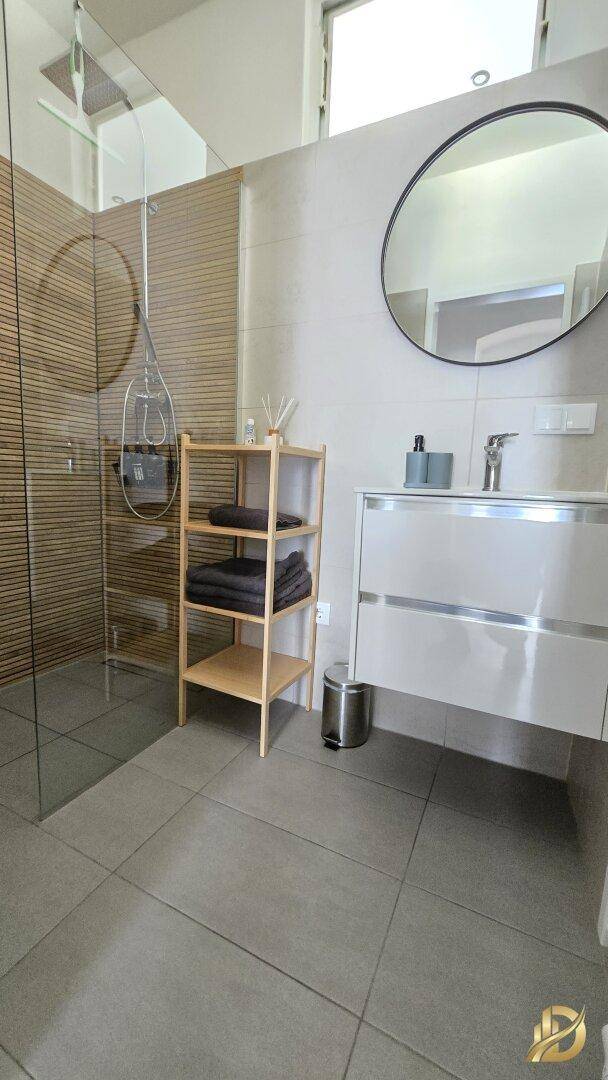 Modernes Badezimmer mit begehbarer Dusche, Waschtisch und rundem Spiegel, in hellen Grautönen gehalten.