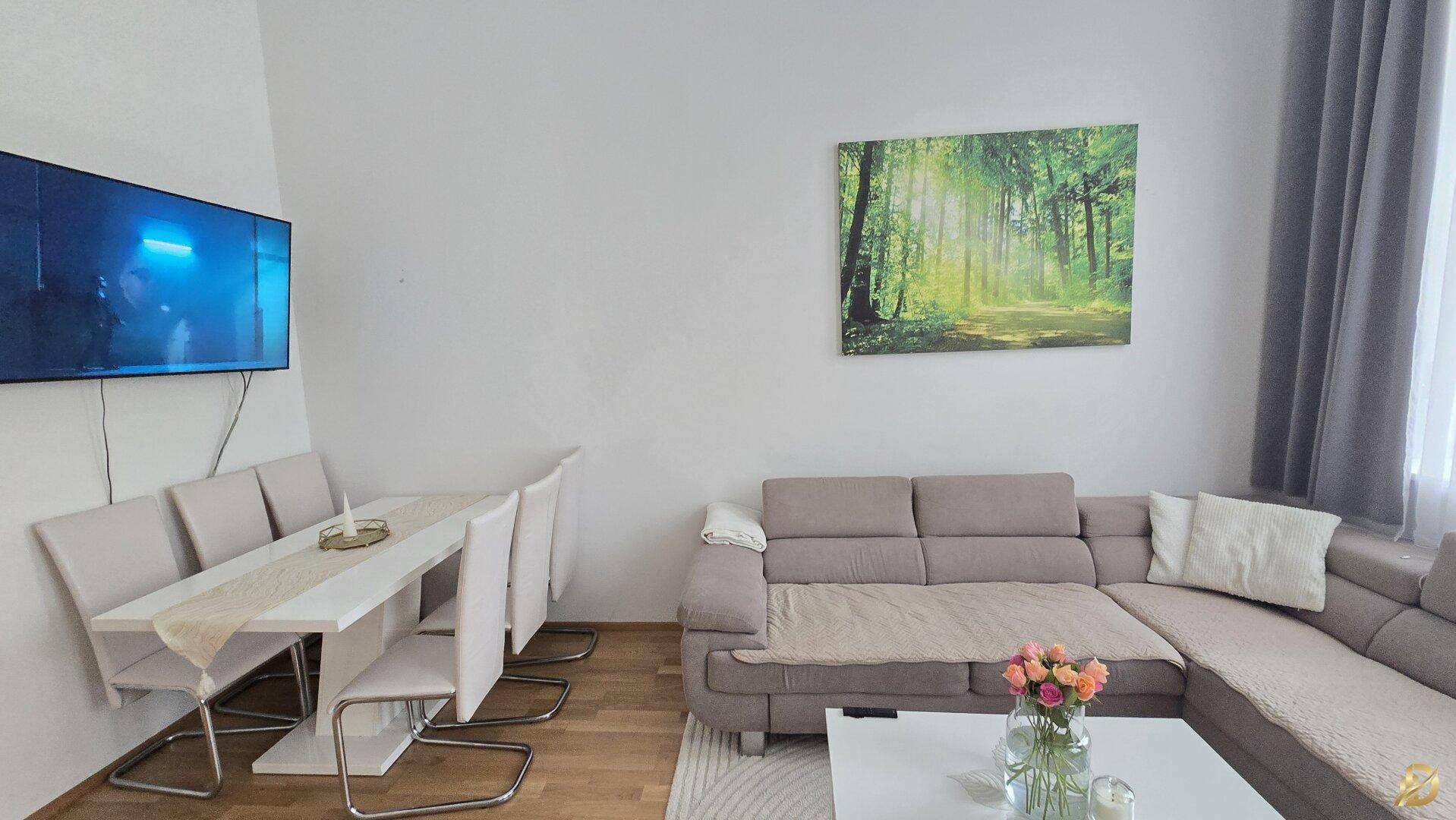 Gemütlicher Wohnbereich mit Ecksofa, Esstisch und Wandfernseher, dekoriert mit einem Waldgemälde.
