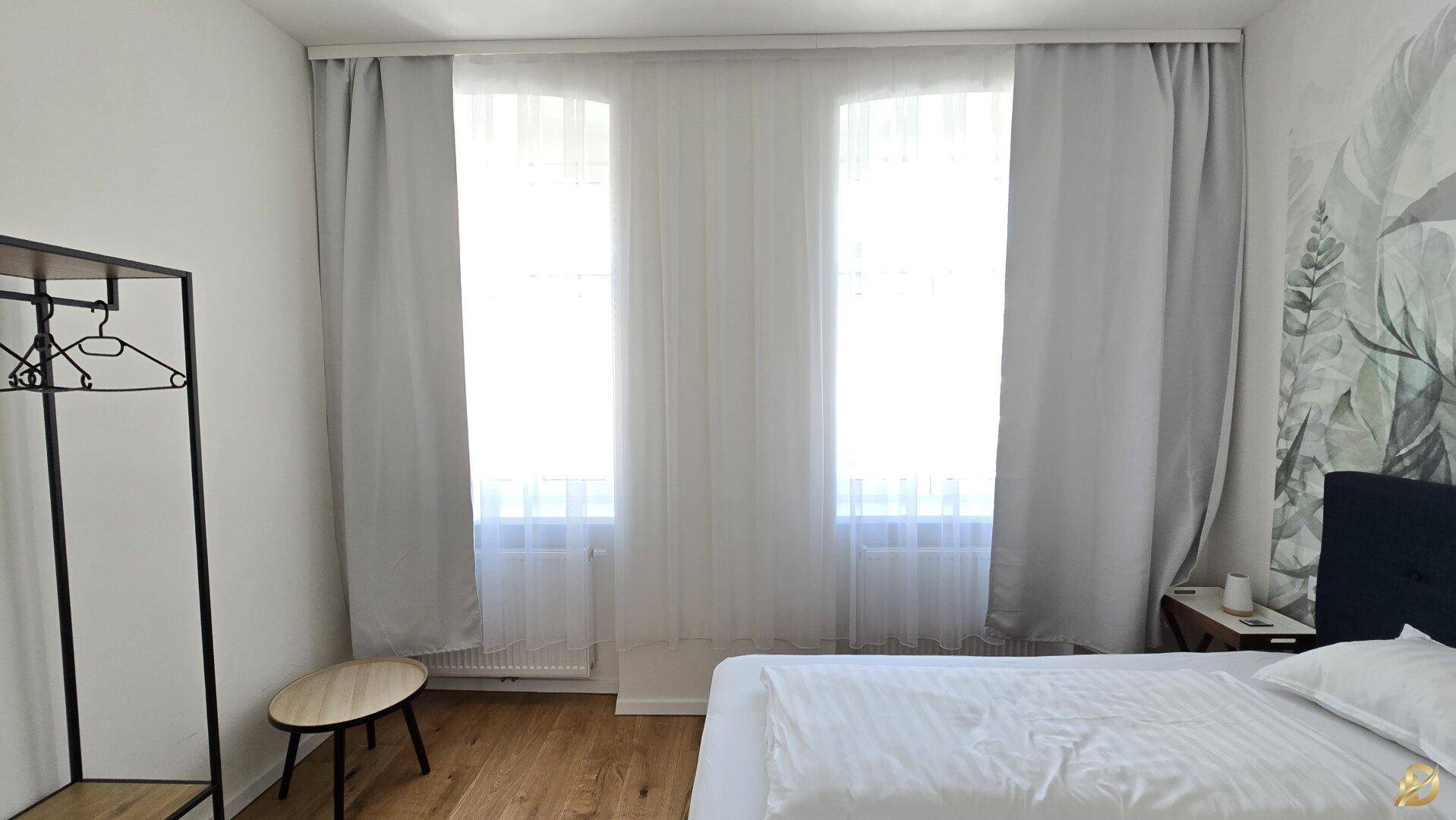 Schlafzimmer mit Holzboden, großen Fenstern und offenem Kleiderständer.