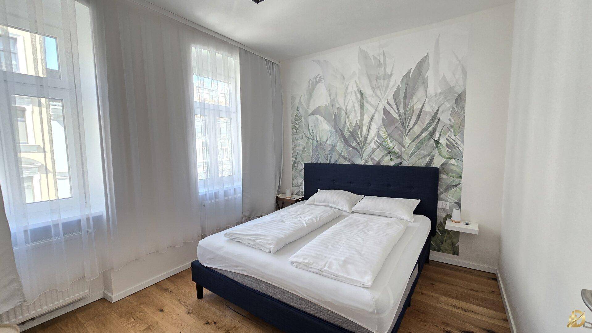 Gemütliches Schlafzimmer mit Doppelbett, großen Fenstern und floraler Wandgestaltung.