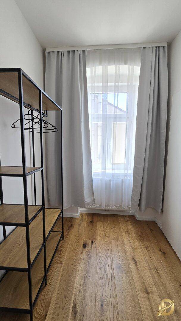 Kleiner Raum mit Holzboden, Fenster und offenem Kleiderständer.