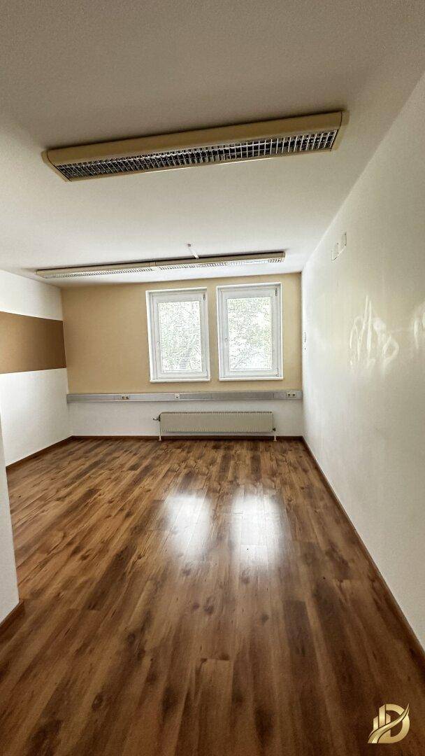 Büro mit Laminatboden, einer beigen Akzentwand und zwei Fenstern, die für natürliche Belichtung sorgen.