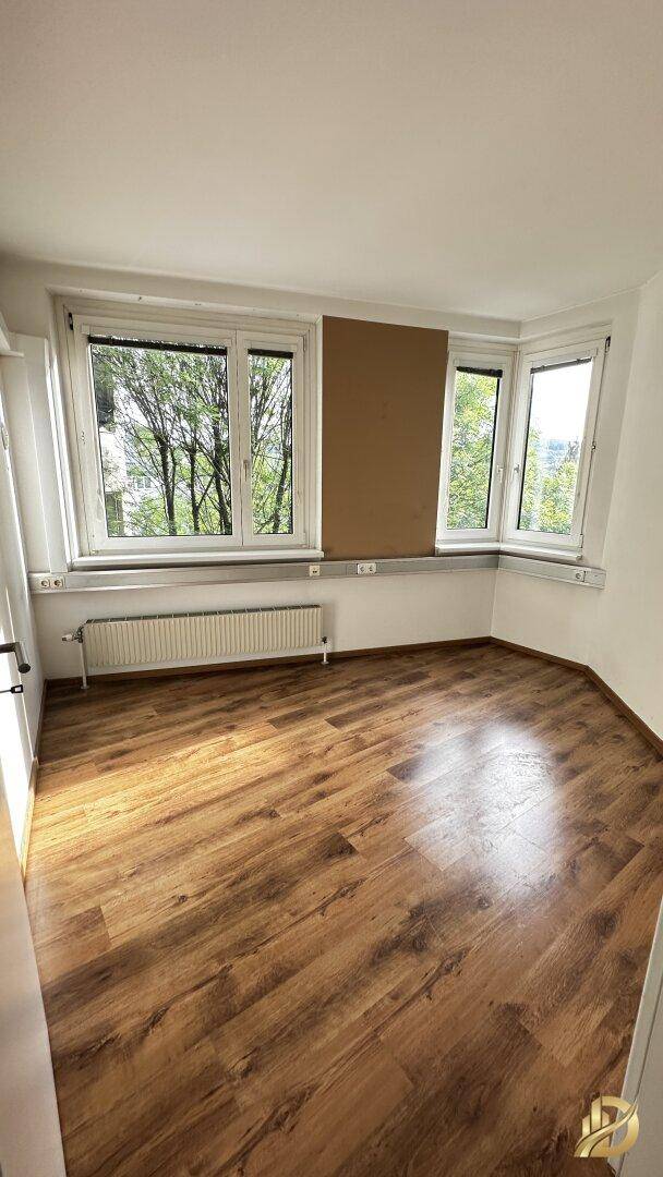 Helles Eckbüro mit Laminatboden und mehreren Fenstern, die einen Blick ins Grüne ermöglichen.