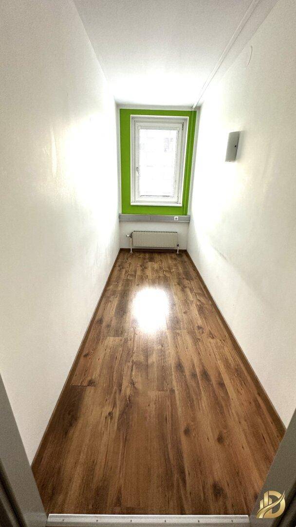 Langer, schmaler Büroraum mit Laminatboden, grüner Akzentwand und einem Fenster am Ende.