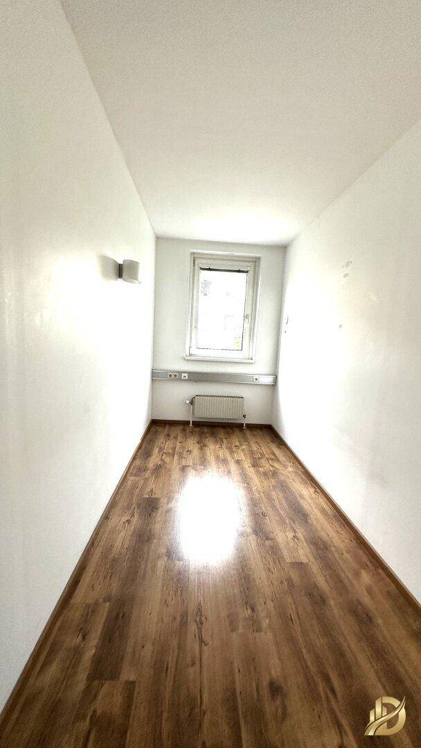 Langer, schmaler Büroraum mit Laminatboden und einem Fenster, das für ausreichend Helligkeit sorgt.