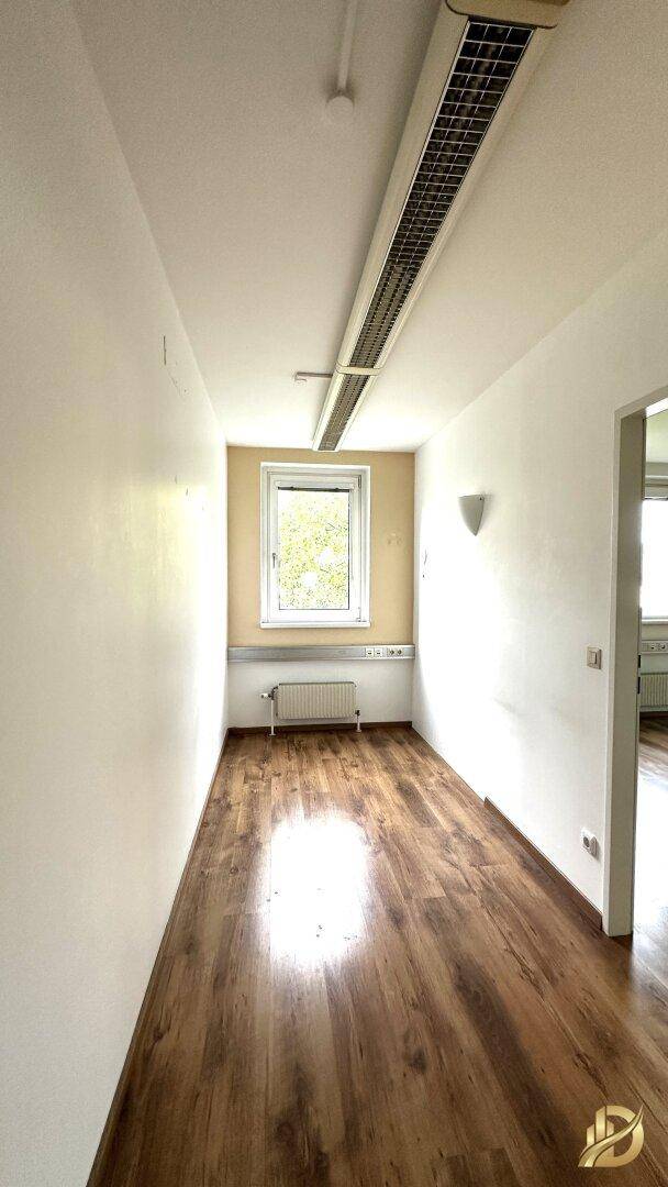 Langer, schmaler Büroraum mit Laminatboden, beiger Akzentwand und einem Fenster am Ende.