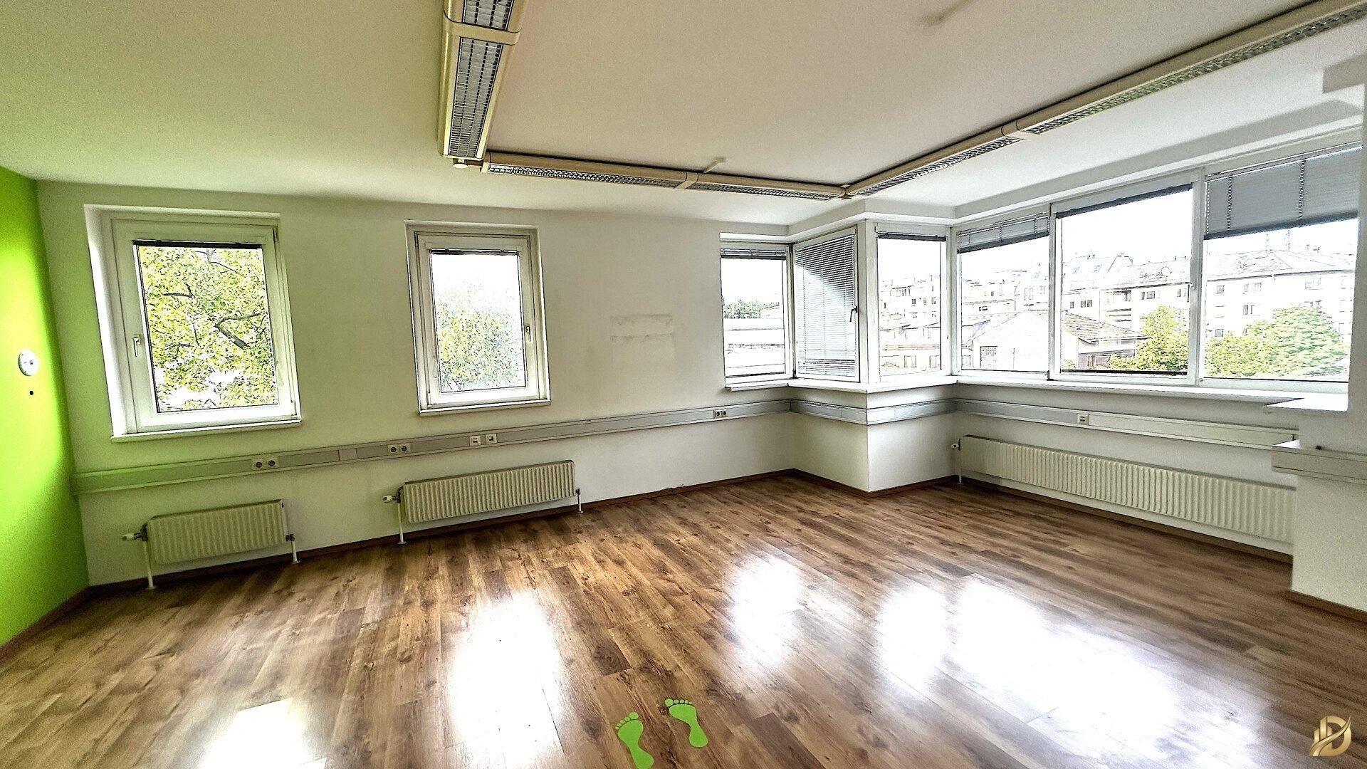 Großes Eckbüro mit Laminatboden, grüner Akzentwand und vielen Fenstern mit Blick ins Grüne.