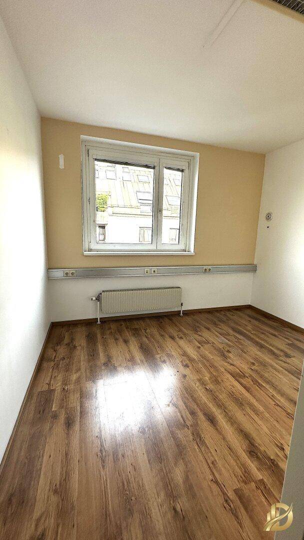 Büro mit Laminatboden, beiger Wand und einem großen Fenster, das den Raum mit Tageslicht füllt.