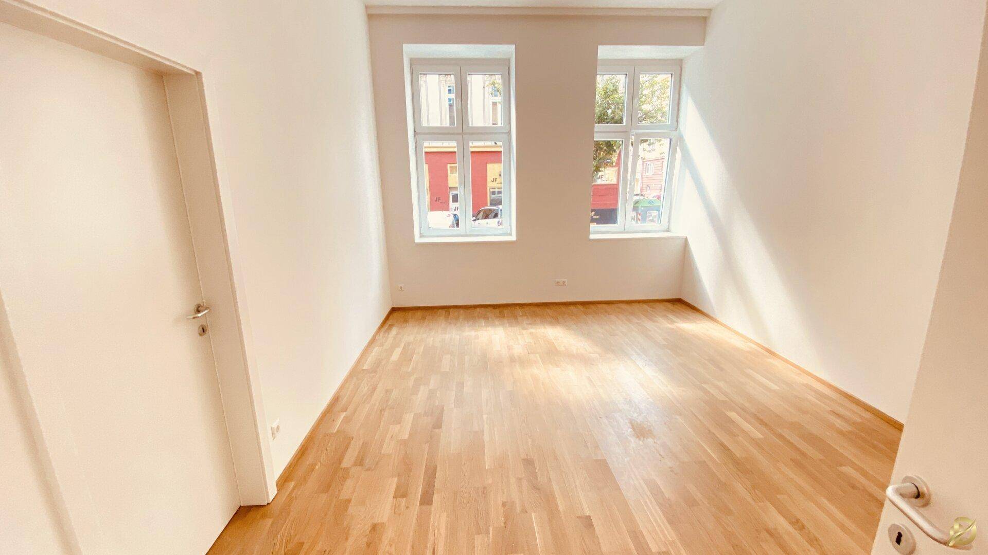 Geräumiges, leeres Zimmer mit Holzboden und zwei großen Fenstern.