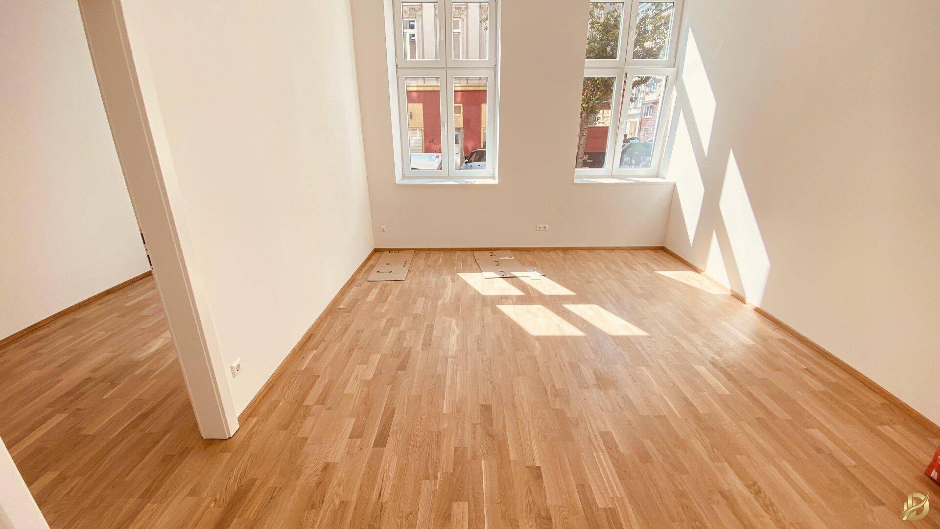 Helles Zimmer mit Holzboden und großen Fenstern, das viel Tageslicht hereinlässt.