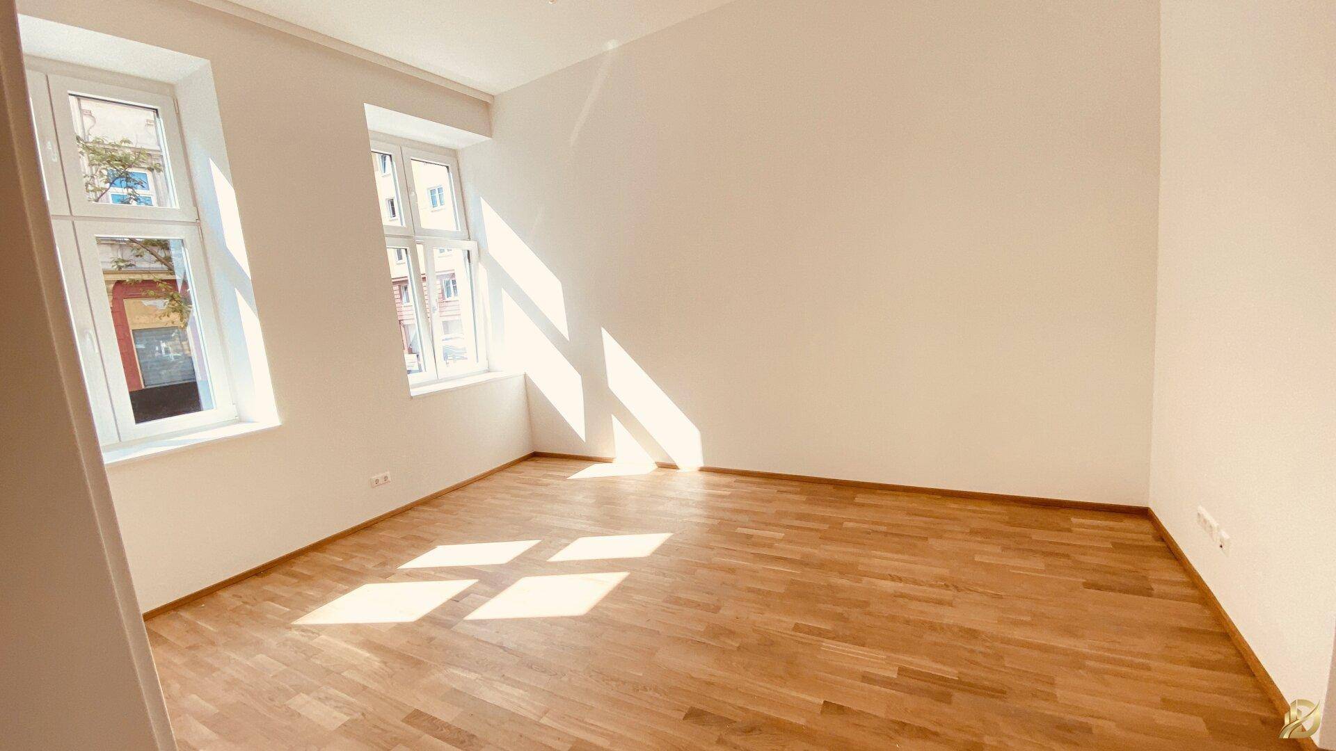 Geräumiges, leeres Zimmer mit Holzboden und großen Fenstern, die viel Sonne hereinlassen.