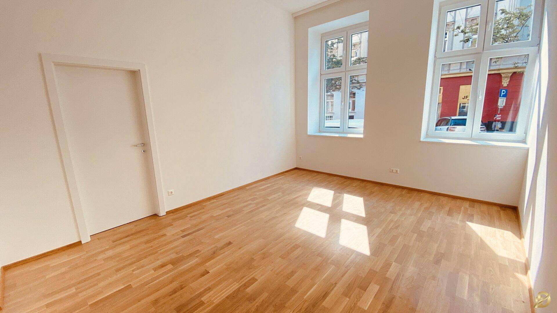 Helles Zimmer mit Holzboden, einer Tür und großen Fenstern mit Blick auf die Straße.