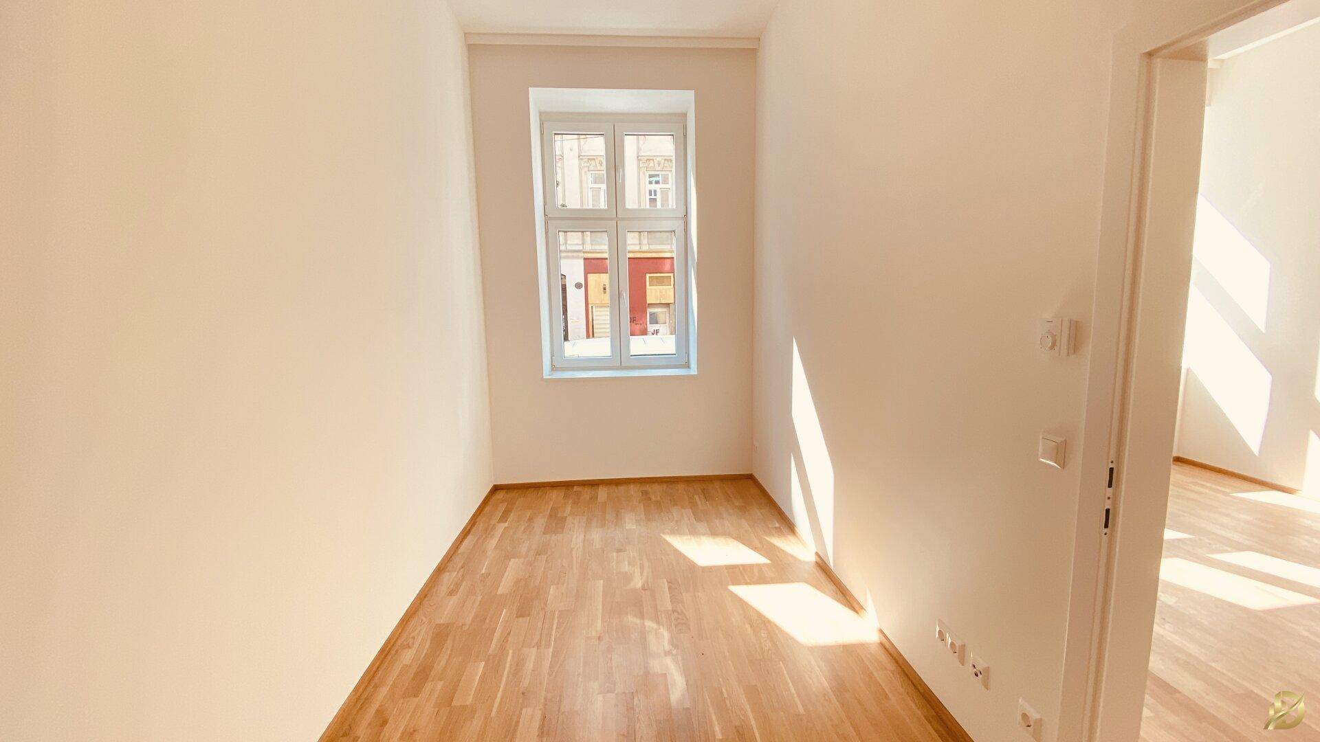 Helles, leeres Zimmer mit Holzboden und einem Fenster, das viel Tageslicht hereinlässt.