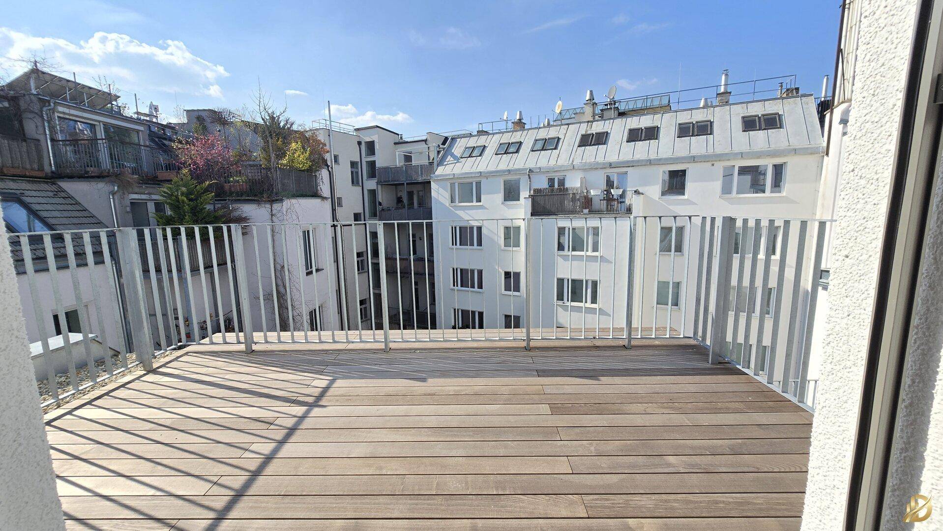 Geräumiger Balkon mit Holzdeck und Metallgeländer, bietet einen Blick auf die umliegenden Gebäude.