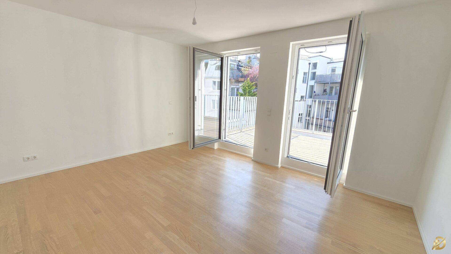 Geräumiges Zimmer mit hellem Parkettboden und großen Fenstern, die auf einen Balkon führen.