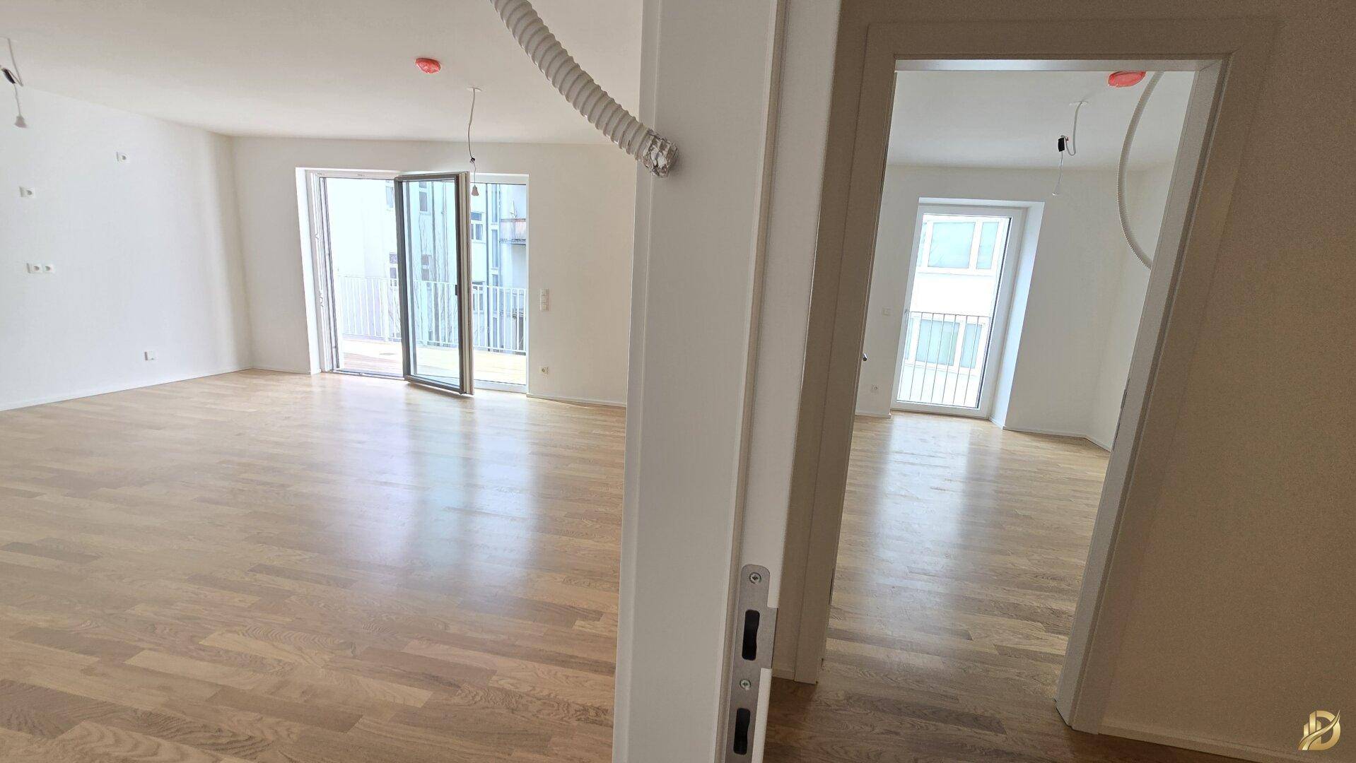 Blick von einem hellen Raum mit Parkettboden in einen weiteren, ähnlich gestalteten Wohnbereich.