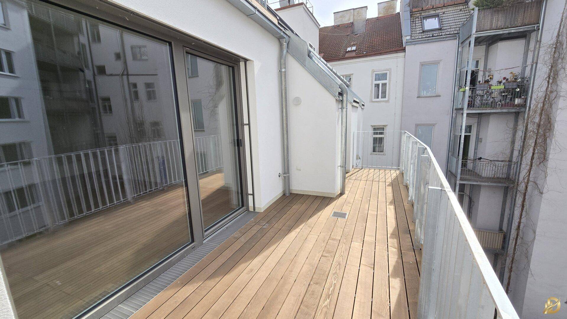 Geräumige Holzterrasse mit Blick auf die umliegenden Gebäude und Glasschiebetüren zum Innenbereich.