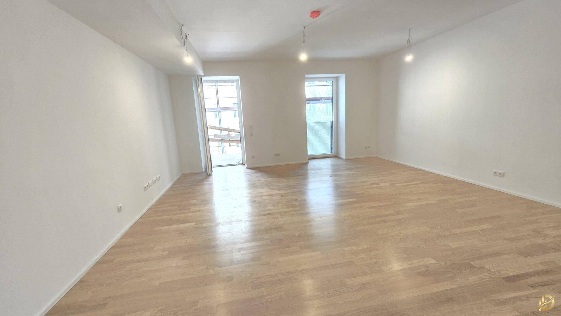 Helles, leeres Wohnzimmer mit Parkettboden und Zugang zu einem Balkon.