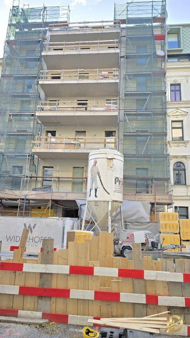 Fassade eines im Bau befindlichen Gebäudes, umgeben von Gerüsten und Baumaterialien auf der Baustelle.