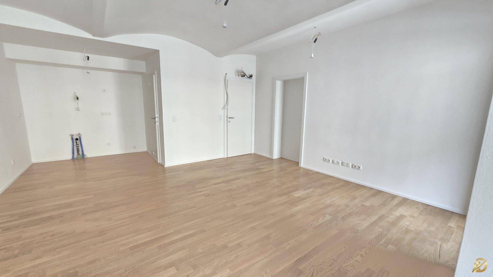 Geräumiger, heller Raum mit Holzparkett und Anschlüssen für eine zukünftige Küchenzeile.