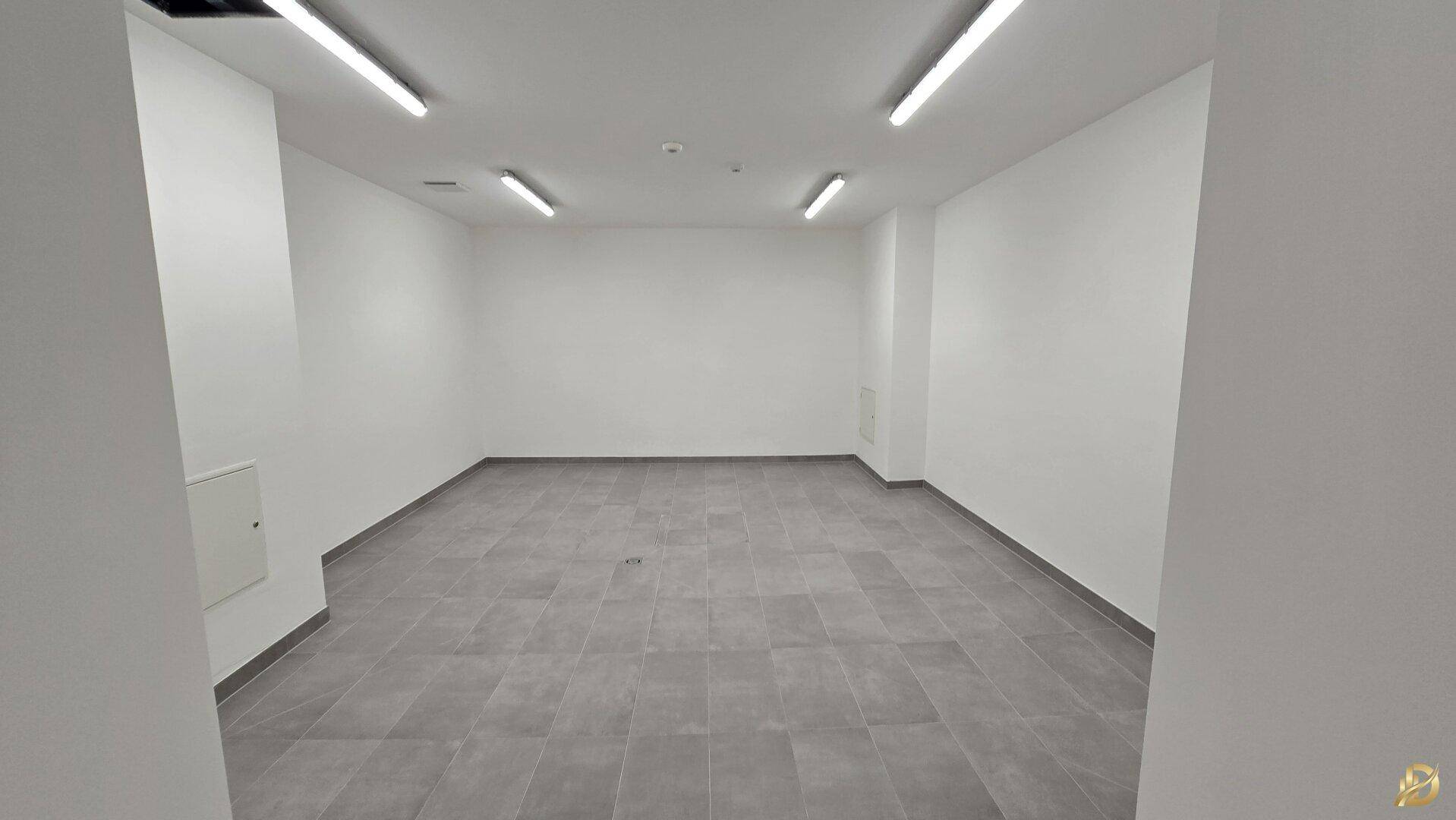 Heller, leerer Raum mit weißen Wänden und grauen Fliesen, beleuchtet von Deckenleuchten.