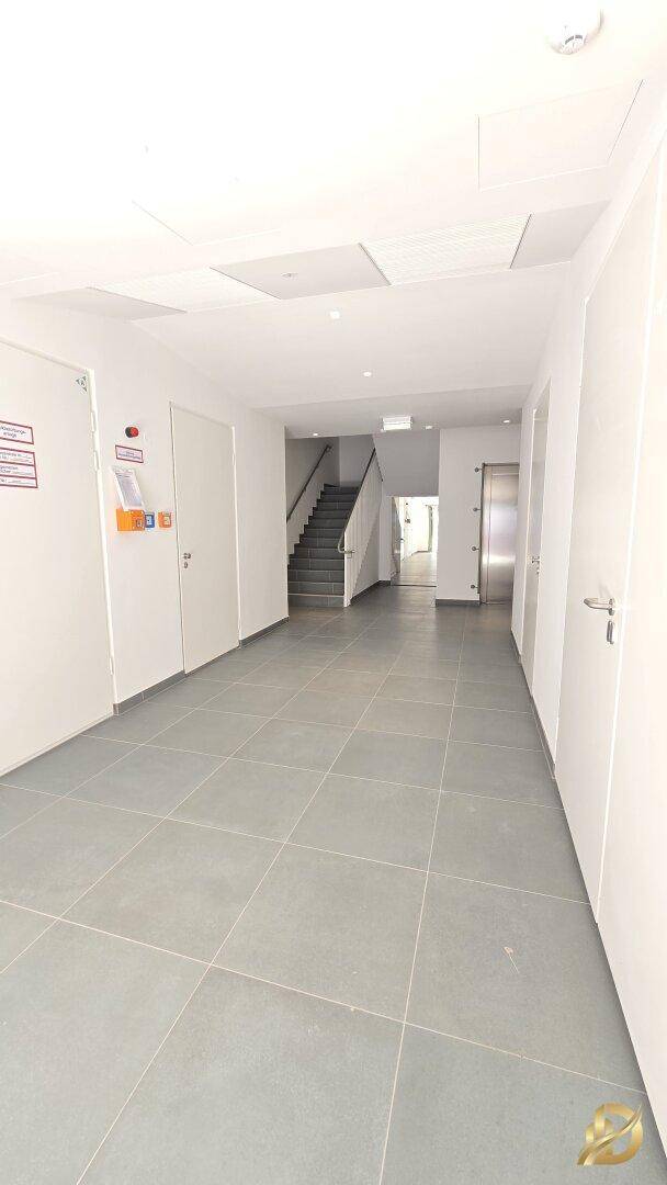 Langer Flur mit mehreren Türen, grauen Fliesen und einem Treppenhaus sowie Aufzug im Hintergrund.