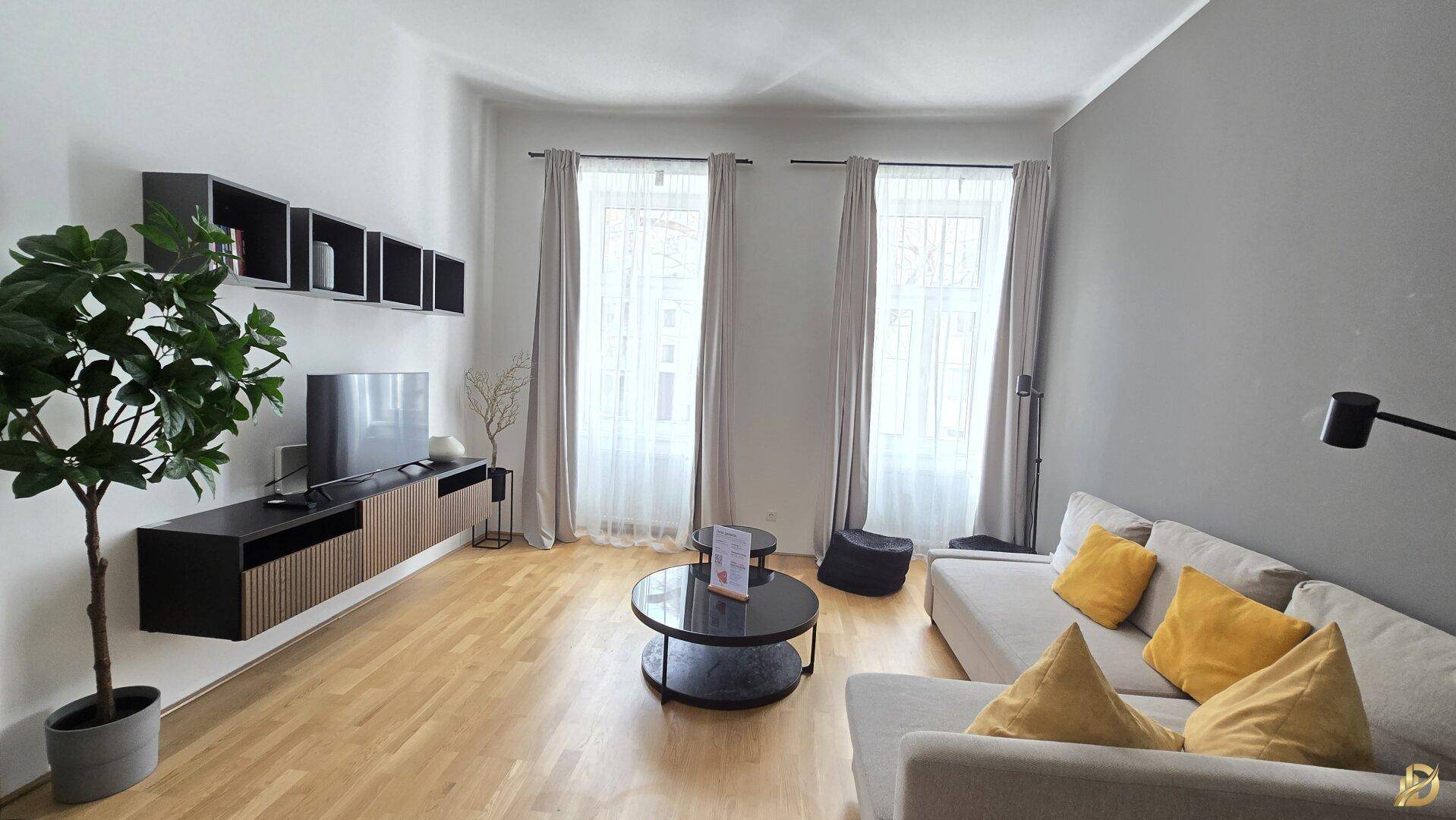 Geräumiges Wohnzimmer mit zwei großen Fenstern, Holzboden und moderner Einrichtung.