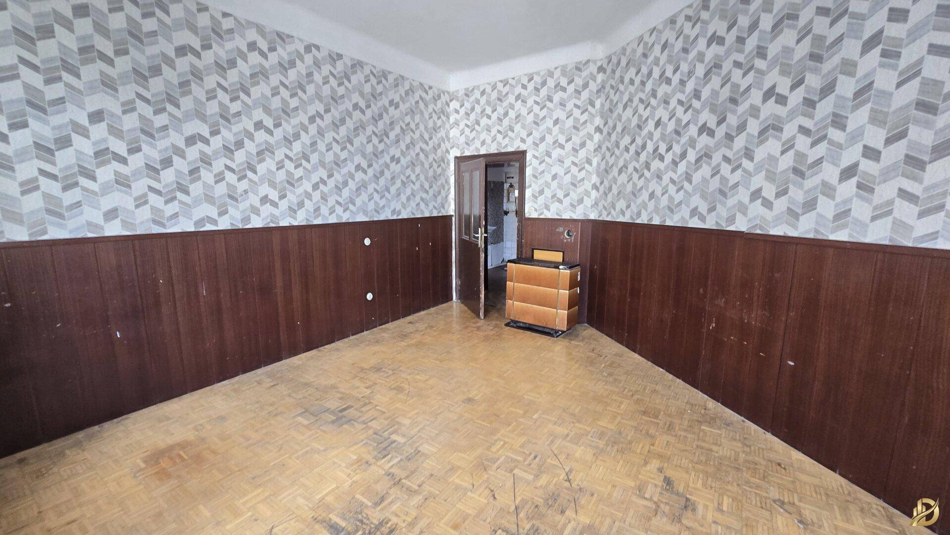 Großes Zimmer mit Fischgrätparkett und gemusterter Tapete, mit Blick auf den Eingangsbereich.