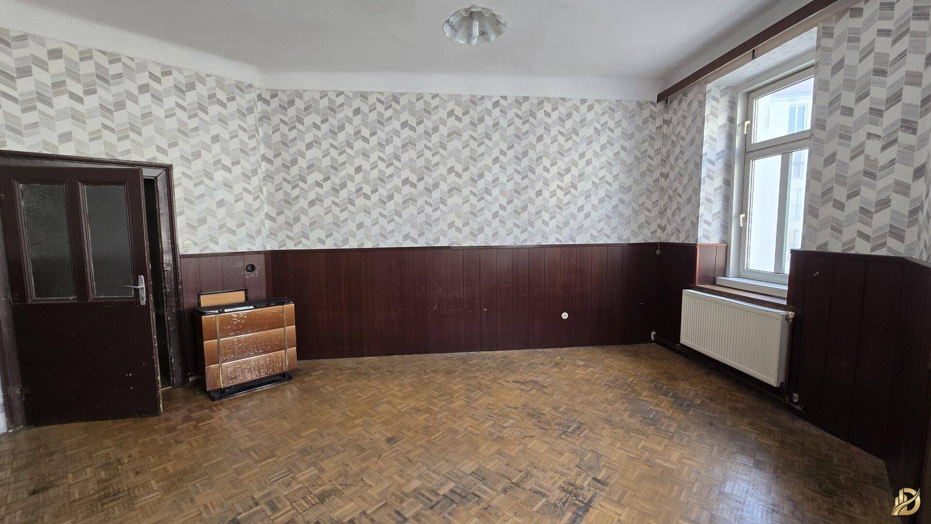 Geräumiges Zimmer mit Fischgrätparkett, Holzvertäfelung und Heizkörper, bereit für eine Renovierung.