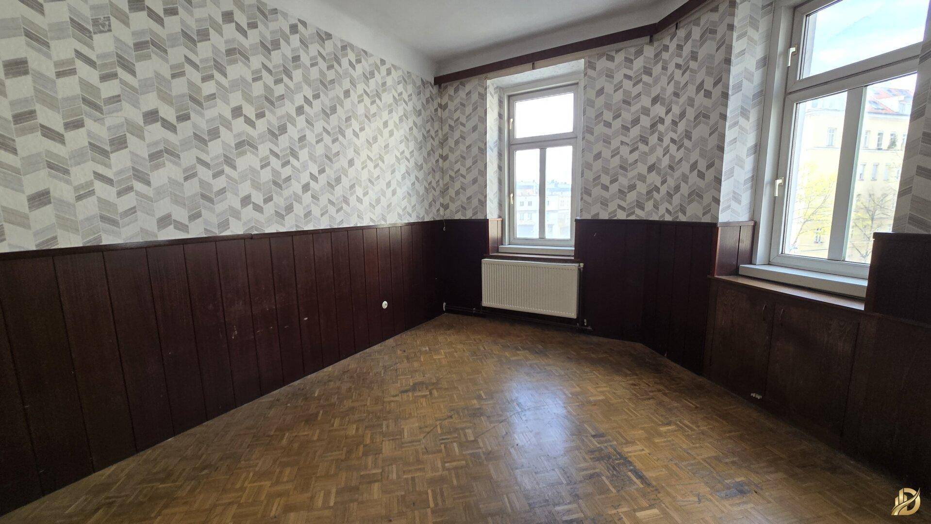 Helles Zimmer mit Fischgrätparkett, gemusterter Tapete und Holzvertäfelung, mit Blick nach draußen.