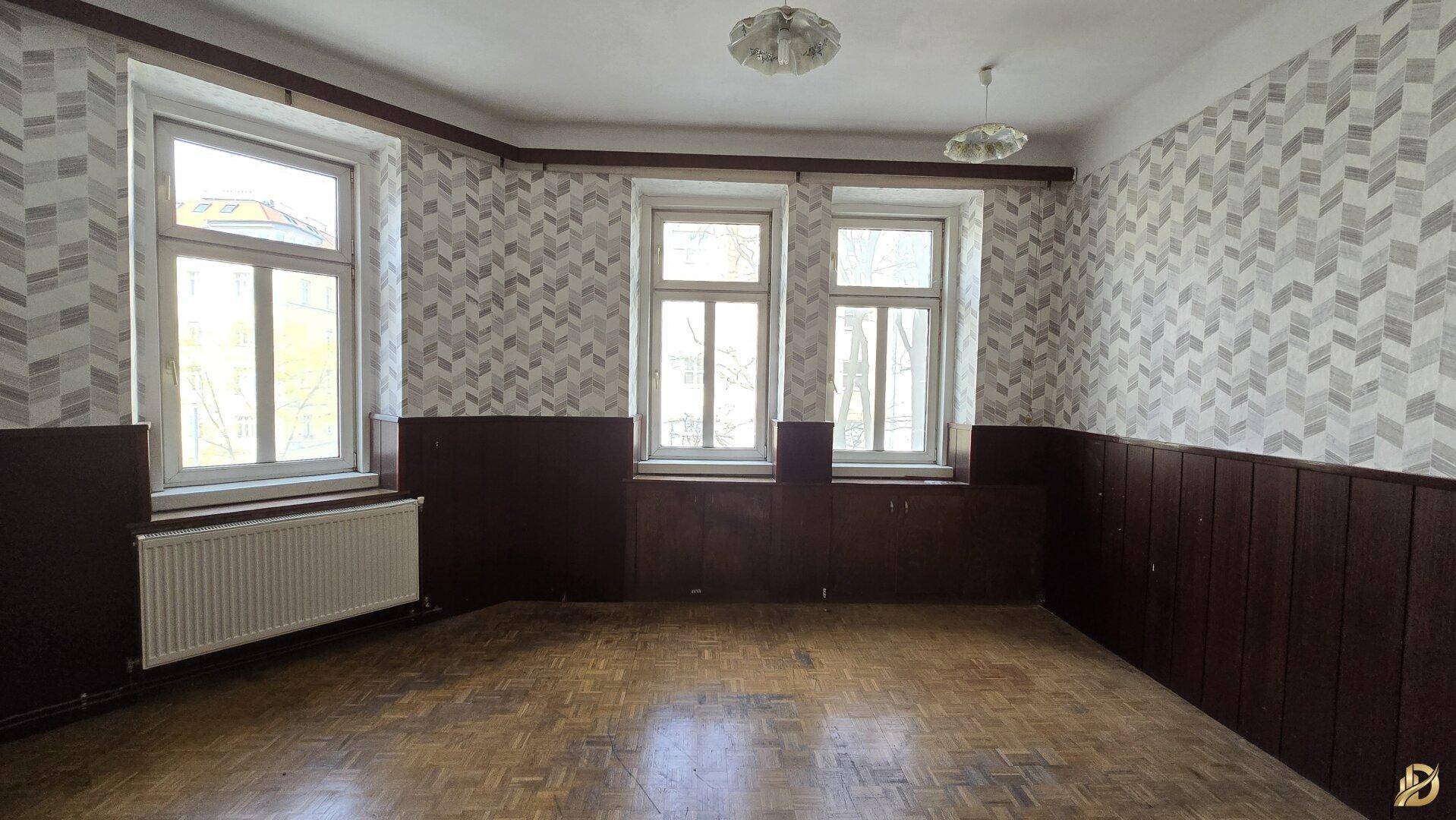 Helles Zimmer mit Fischgrätparkett, gemusterter Tapete und Holzvertäfelung, ideal für eine Neugestaltung.