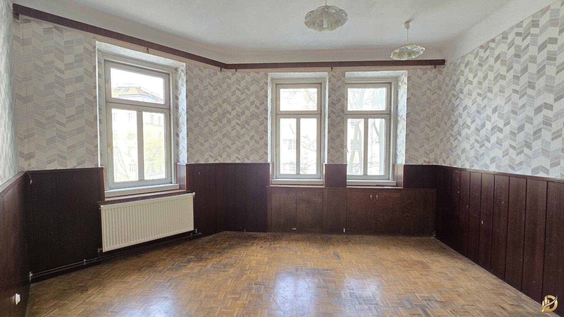 Großes Zimmer mit Fischgrätparkett, Holzvertäfelung und drei Fenstern, wartet auf eine Modernisierung.