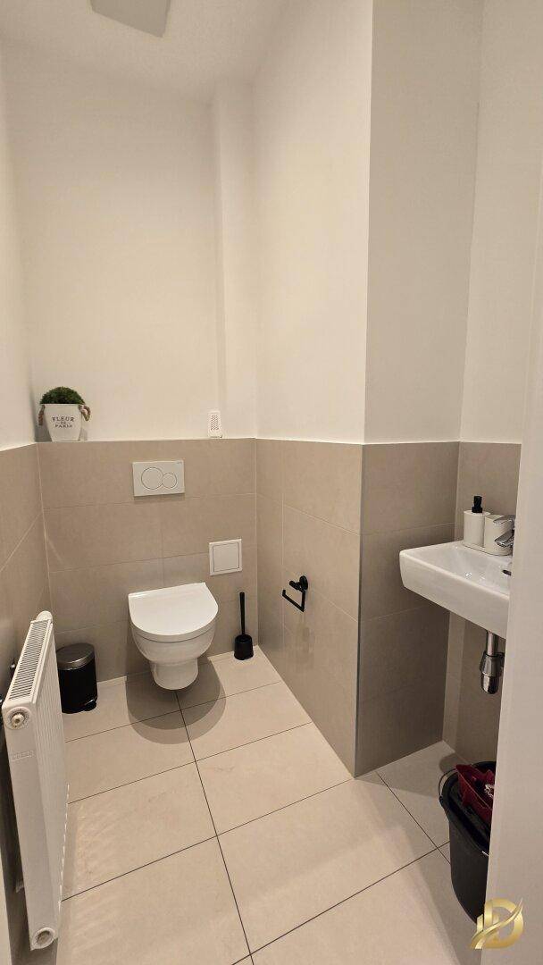 Separates WC mit wandhängender Toilette und kleinem Waschbecken, helle Fliesen.