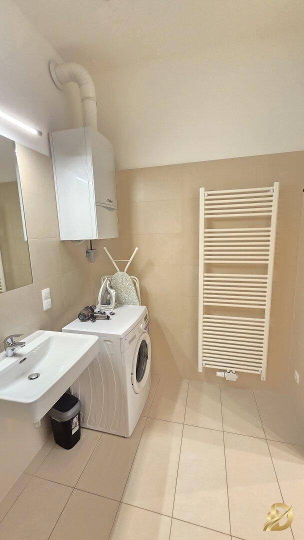 Badezimmer mit Waschmaschine, Waschbecken und beheiztem Handtuchhalter, helle Fliesen.